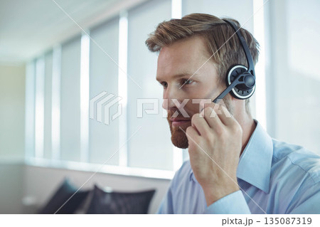 Male office worker wearing dress shirt adjusting headset mic at office desk with blinds, copy space Male office worker wearing dress shirt adjusting headset mic at office desk with blinds, copy space 135087319