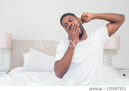 African American man yawning and stretching in bedroom during morning beside king-size bed African American man yawning and stretching in bedroom during morning beside king-size bed 135087323