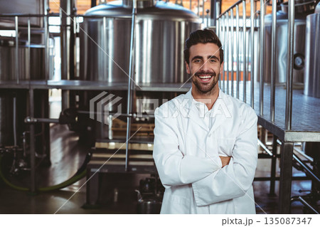 Male brewery technician wearing lab coat crossing arms on brewery floor near fermentation tanks Male brewery technician wearing lab coat crossing arms on brewery floor near fermentation tanks 135087347