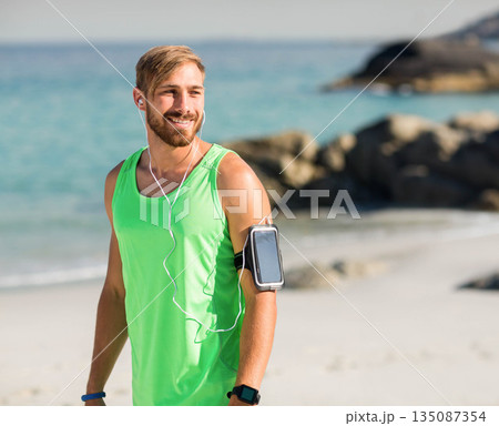 Man wearing tank top, watch, smartphone armband, wired earphones pausing run on beach, copy space 135087354