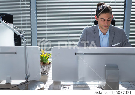Male professional working on dual computer monitors at office desk with succulent plant and lamp Male professional working on dual computer monitors at office desk with succulent plant and lamp 135087371