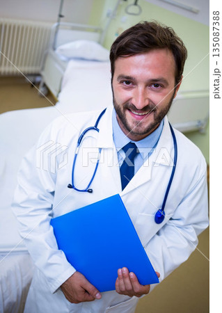 Male doctor standing in patient room holding blue file folder with stethoscope beside hospital bed Male doctor standing in patient room holding blue file folder with stethoscope beside hospital bed 135087388