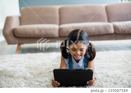 Girl holding black tablet lying on white shag carpet in living room with brown sofa Girl holding black tablet lying on white shag carpet in living room with brown sofa 135087389
