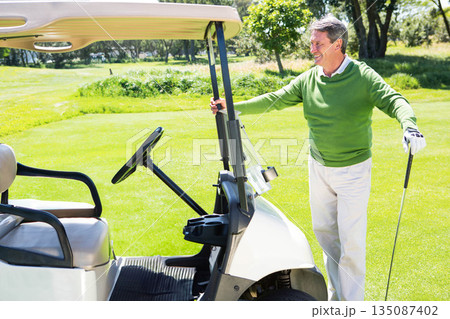 Senior African American man smiling while holding golf club beside golf cart on fairway, copy space 135087402