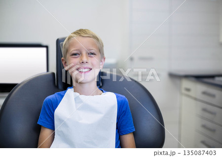 Child patient sitting on reclining dental chair in treatment room wearing protective bib by monitor 135087403