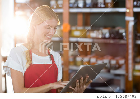 Female store clerk standing by wooden shelves at boutique shop holding tablet checking glass jars Female store clerk standing by wooden shelves at boutique shop holding tablet checking glass jars 135087405