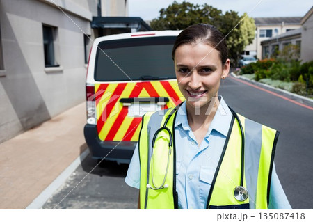 Female EMT standing outside clinic with chevron emergency vehicle, vest and stethoscope, copy space 135087418