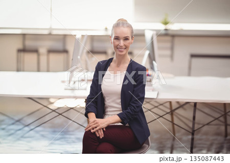 Businesswoman sitting on swivel chair in modern office working at dual monitors wearing navy blazer 135087443