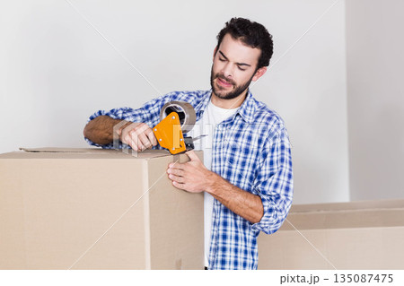 Man using tape dispenser sealing large cardboard box in empty room with packed box behind 135087475
