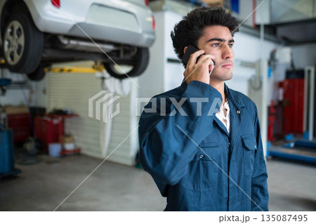 Man in twenties in coverall holding smartphone to ear and checking hatchback at garage, copy space 135087495