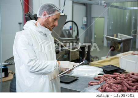 Man wearing white coat hairnet reviewing clipboard at meat plant near raw sausages and machinery Man wearing white coat hairnet reviewing clipboard at meat plant near raw sausages and machinery 135087502