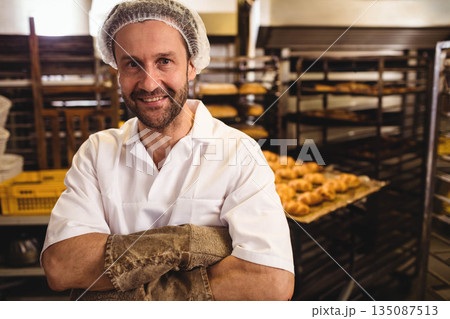 Male baker standing in bakery area wearing hairnet, leather oven gloves before metal racks of buns Male baker standing in bakery area wearing hairnet, leather oven gloves before metal racks of buns 135087513