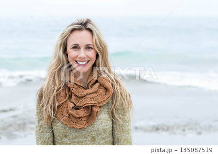 Woman standing on beach smiling at camera wearing olive green sweater with textured brown scarf 135087519