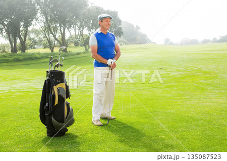 Senior man wearing sleeveless sweater and glove while holding golf club on fairway near golf bag Senior man wearing sleeveless sweater and glove while holding golf club on fairway near golf bag 135087523