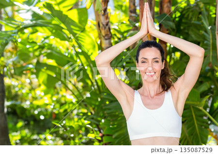 Woman raising arms in yoga prayer pose outdoors in tropical garden wearing sports bra, copy space Woman raising arms in yoga prayer pose outdoors in tropical garden wearing sports bra, copy space 135087529