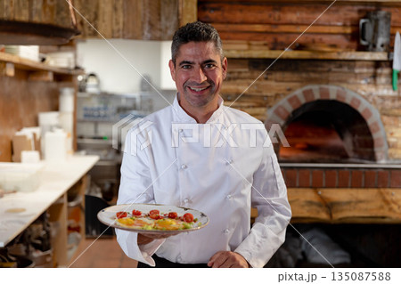 Mature male chef in white jacket presenting appetizer plate at prep counter near wood-fired oven Mature male chef in white jacket presenting appetizer plate at prep counter near wood-fired oven 135087588