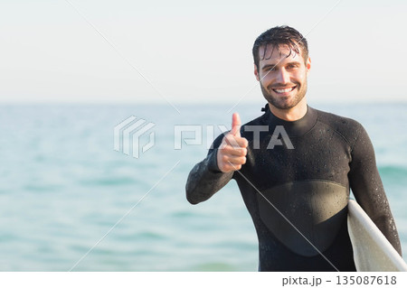 Man standing at shoreline holding white surfboard and wearing black wetsuit by ocean, copy space Man standing at shoreline holding white surfboard and wearing black wetsuit by ocean, copy space 135087618