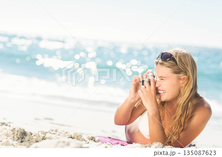 Woman lying on white sand at shoreline wearing bikini top and holding compact camera, copy space 135087623