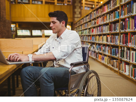 Man in wheelchair typing on laptop at wooden study table inside library with bookshelves Man in wheelchair typing on laptop at wooden study table inside library with bookshelves 135087624