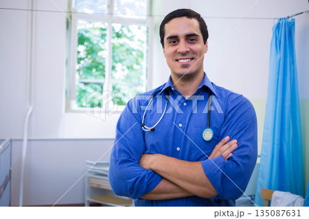 Middle-aged man standing with arms crossed in medical exam room wearing stethoscope by window Middle-aged man standing with arms crossed in medical exam room wearing stethoscope by window 135087631
