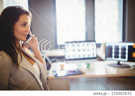 Woman wearing blazer standing by office desk with monitors holding smartphone to ear, copy space 135087635