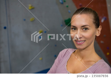 Woman standing with arms folded facing camera at climbing gym with padded floor and colorful holds 135087656
