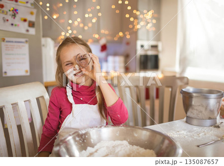 Child girl holding cookie cutter over eye mixing flour in bowl on table at home kitchen Child girl holding cookie cutter over eye mixing flour in bowl on table at home kitchen 135087657