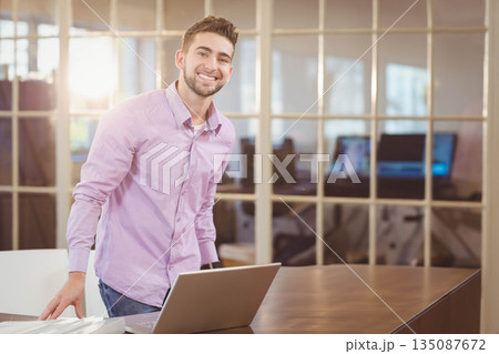 Male professional leaning on office table beside laptop, papers behind glass partition, copy space Male professional leaning on office table beside laptop, papers behind glass partition, copy space 135087672