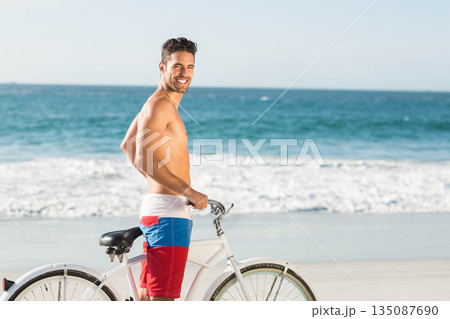 Shirtless man wearing red white blue trunks smiling on beach next to cruiser bicycle, glancing back 135087690