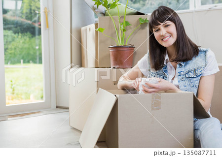 Woman wearing vest sitting on floor in room unpacking moving boxes with bubble wrap, copy space 135087711