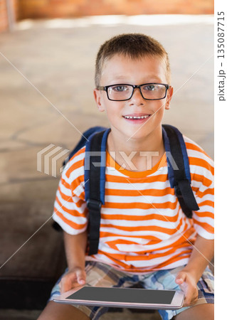 School-aged boy with eyeglasses sitting on tiled step in covered patio holding tablet with backpack School-aged boy with eyeglasses sitting on tiled step in covered patio holding tablet with backpack 135087715