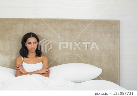 African American woman sitting on bed in bedroom, crossing arms before beige headboard and pillows African American woman sitting on bed in bedroom, crossing arms before beige headboard and pillows 135087731