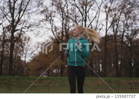 Woman jogging on grassy field at park wearing teal zip-up hoodie and gray gloves Woman jogging on grassy field at park wearing teal zip-up hoodie and gray gloves 135087737