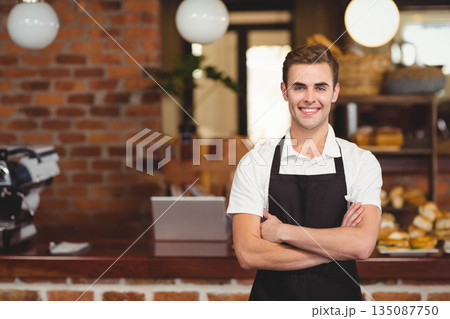 Male barista wearing apron behind cafe counter crossing arms near espresso machine, copy space 135087750