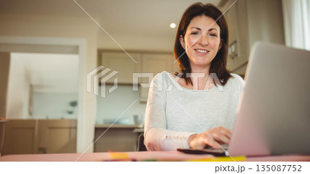 Woman typing on laptop at bright kitchen workspace with light wood cabinets behind, copy space Woman typing on laptop at bright kitchen workspace with light wood cabinets behind, copy space 135087752