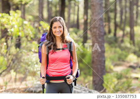 Woman hiker trekking on pine woodland trail wearing athletic gear and carrying backpack and poles 135087753