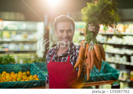 Male grocery worker wearing red apron holding bunch of carrots toward camera in produce aisle 135087762