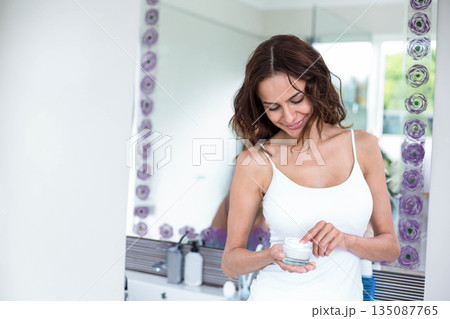 Woman wearing white camisole standing at bathroom vanity applying face cream from jar, copy space 135087765