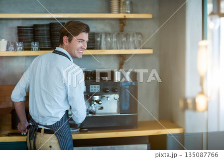 Male barista operating commercial espresso machine behind counter by shelves stacked with cups 135087766