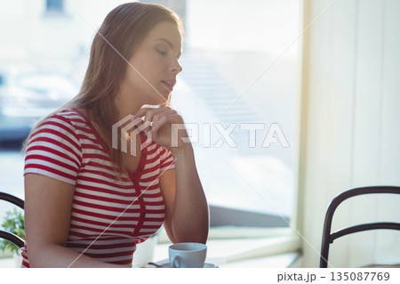 Woman sitting at cafe table by window resting chin on hand gazing over white coffee cup 135087769