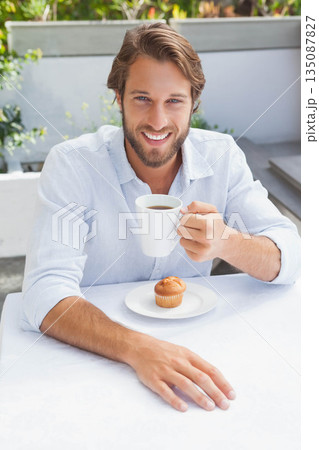 Man smiling while sitting at outdoor cafe terrace on white tablecloth holding coffee mug and muffin Man smiling while sitting at outdoor cafe terrace on white tablecloth holding coffee mug and muffin 135087827