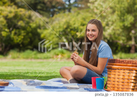 Child female sitting on blanket in park holding sandwich near picnic basket and cups, copy space Child female sitting on blanket in park holding sandwich near picnic basket and cups, copy space 135087836