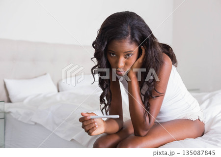 African American woman in sleepwear sitting on bed holding pregnancy test stick looking concerned African American woman in sleepwear sitting on bed holding pregnancy test stick looking concerned 135087845