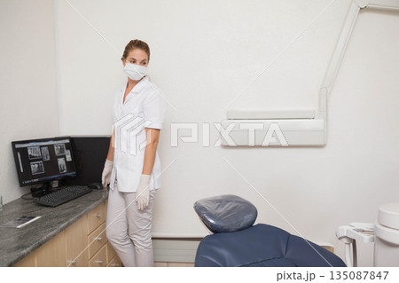 Female dentist wearing mask standing by countertop in clinic viewing X-rays on monitor, copy space 135087847