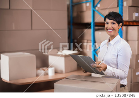 Woman holding clipboard and pen checking inventory in warehouse with boxes, tape, copy space 135087849