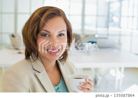 Woman wearing blazer holding white coffee cup smiling at office desk with laptop, copy space Woman wearing blazer holding white coffee cup smiling at office desk with laptop, copy space 135087864