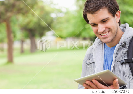 Man standing in grassy park holding tablet with both hands and carrying black backpack, copy space Man standing in grassy park holding tablet with both hands and carrying black backpack, copy space 135087877