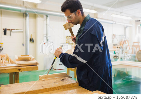 Male woodworker wearing goggles and coverall carving wood block at workbench with mallet and chisel 135087878