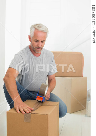 Middle-aged man kneeling in hallway sealing cardboard boxes with tape dispenser, clear packing tape 135087881