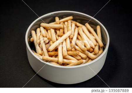 Crispy small breadsticks served on a white glass plate against a black background. Minimalistic food composition highlighting texture, contrast, and simple snack presentation. Crispy small breadsticks served on a white glass plate against a black background. Minimalistic food composition highlighting texture, contrast, and simple snack presentation. 135087889
